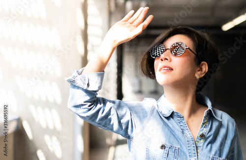 Stylish, middle aged Asian woman wearing 100% UV light protection sunglasses, stand inside and raise her hand to block out bright glare and sunlight from outside to avoid ultraviolet rays overexposure