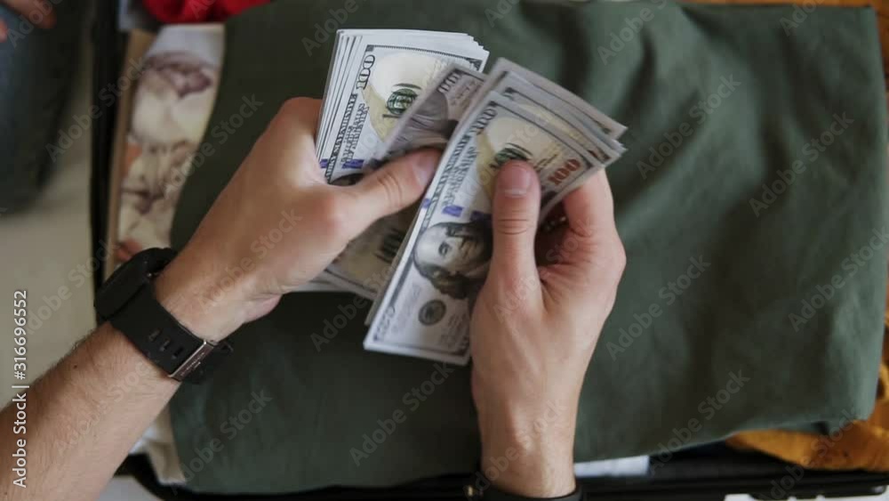 Unrecognizable man hands with pile of money, US dollars notes - counting under the suitcase with clothes. Counting money for holidays, vacation. Top view, close up. Wearing black hand watches