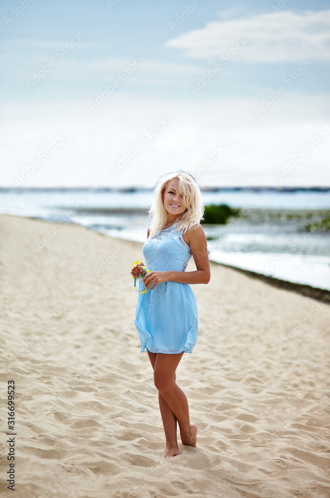 Happy attractive girl relaxing by the sea. Young beautiful woman in blue dress posing outdoors. Female resting at beach in summer. Caucasian girl enjoys a bright summer lifestyle Happy attractive girl relaxing by the sea. Young beautiful woman in blue dress posing outdoors. Female resting at beach in summer. Caucasian girl enjoys a bright summer lifestyle