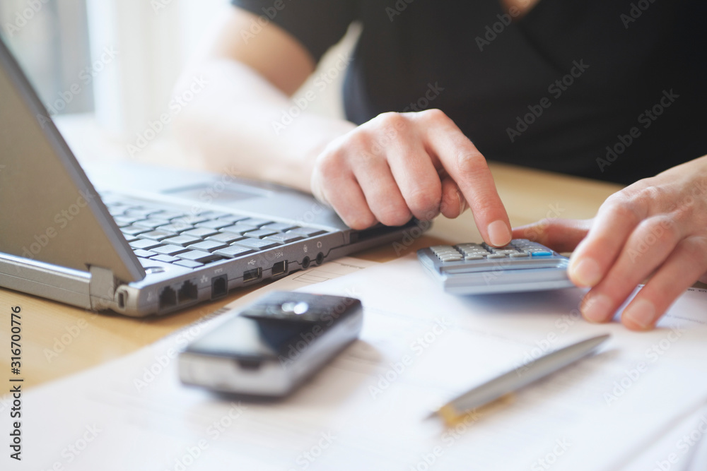 Man using calculator at desk with laptop cell phone and pen in foreground close-up mid section Man using calculator at desk with laptop cell phone and pen in foreground close-up mid section