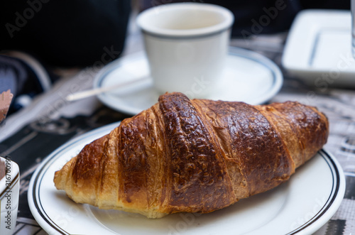 A well-done croissant on white dish in close up view. Blur cup on a saucer background.