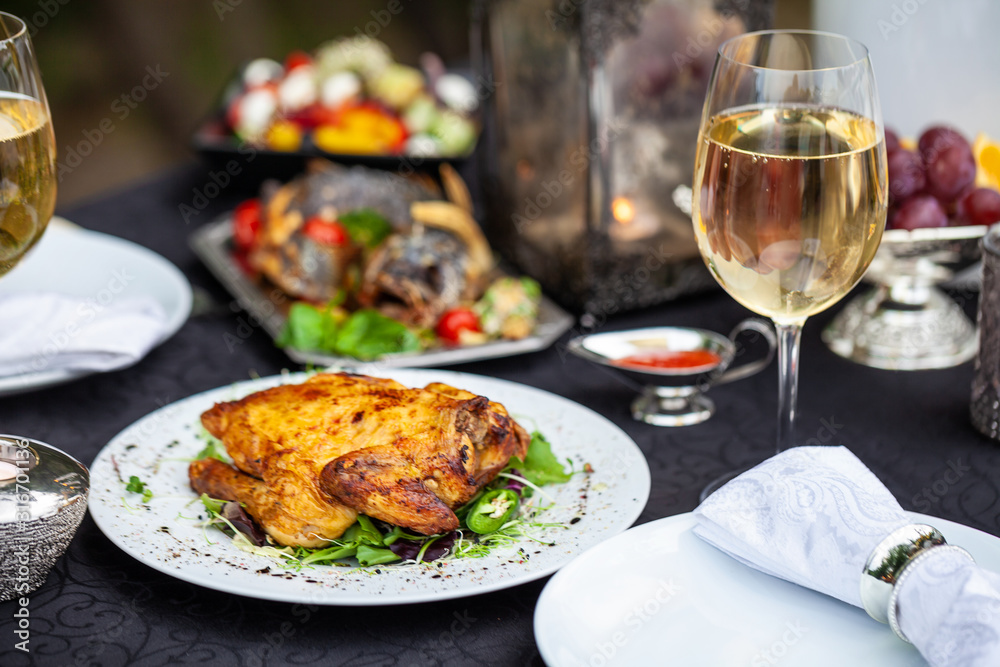 Baked full chicken with vegetables in a plate on a table next to glass of wine and candles. Outdoor, green park Baked full chicken with vegetables in a plate on a table next to glass of wine and candles. Outdoor, green park