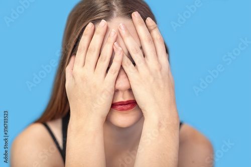 Close up portrait of young caucasian woman covering her face with hands. Bright red lipstick, dark hair, black underwear. Studio, isolated, blue background..