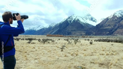 Young adventure photographer alone taking photographs around Mt Cook region, New Zealand. Drone shot tracks forward past man with mountains in background as he aims and focusses his camera. photo