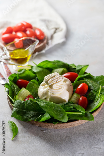 Italian cheese burrata with a salad of fresh spinach, cucumbers and cherry tomatoes with olive oil in a ceramic plate on a light background.