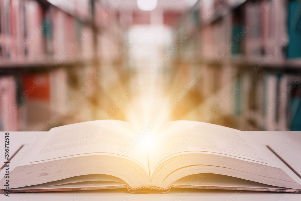 Rays of light coming from an open book or textbook on wooden table with blurred bookshelf in the library. Success education learning or back to school concept background Rays of light coming from an open book or textbook on wooden table with blurred bookshelf in the library. Success education learning or back to school concept background