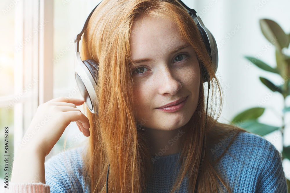 Fototapeta premium pretty smiling young red haired girl with headphones listening to music at home, relaxing, enjoying songs