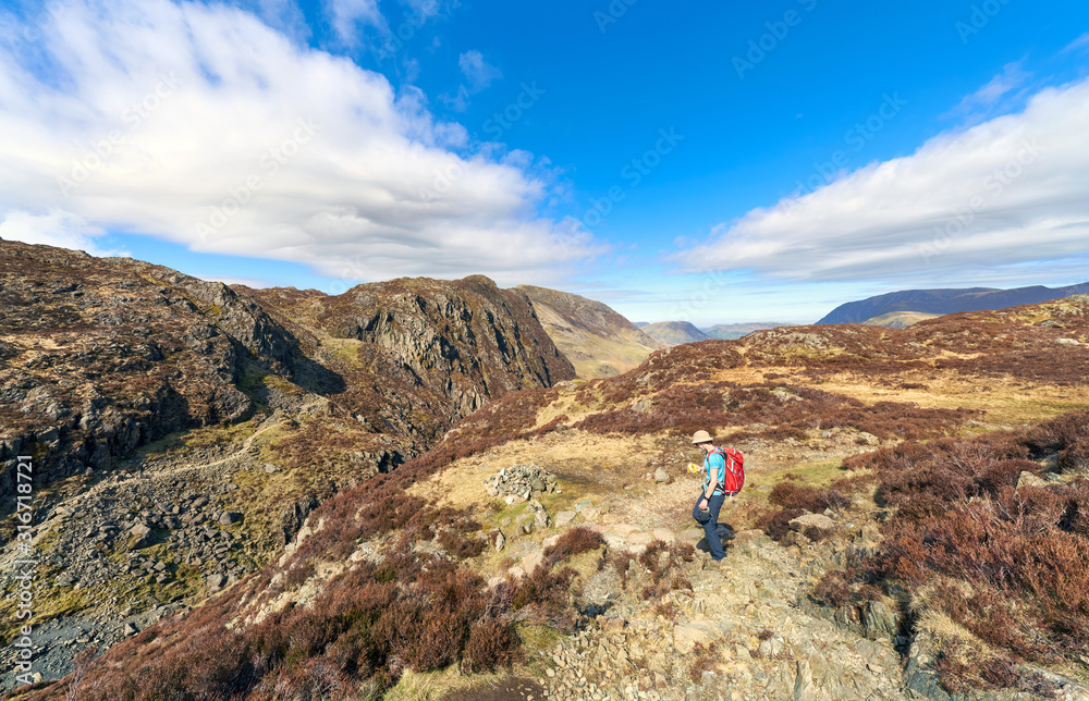 A hiker descending Green Crag with Hay Stacks in the distance on the left on a sunny spring day in the Lake District.