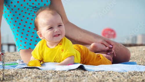 Cheerful cute baby with mom on sandy beach in summer. Happy adorable infant laying on blanket next to sitting mom