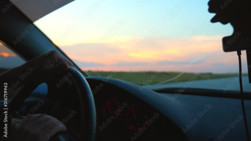 Unrecognizable man holding his hands on steering wheel of car and riding through countryside. Guy enjoying travel journey at evening time. Scenic view on road from inside auto. Slow motion Close up Unrecognizable man holding his hands on steering wheel of car and riding through countryside. Guy enjoying travel journey at evening time. Scenic view on road from inside auto. Slow motion Close up