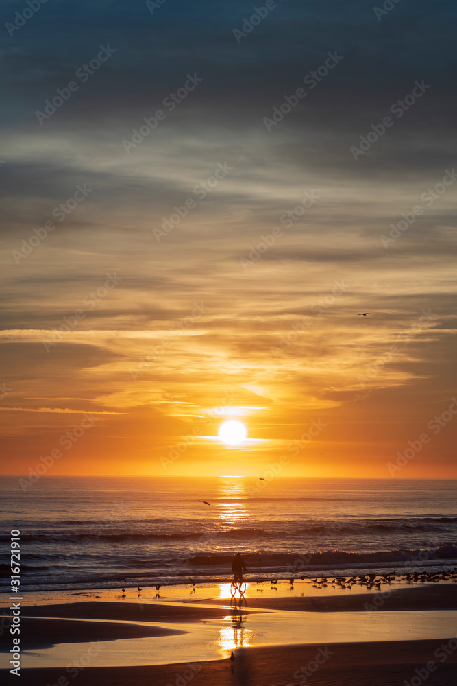 Vertical image of a sunrise along the Atlantic Coast of Port Orange, Florida, featuring a silhouetted man on a bicycle riding on a the beach. 