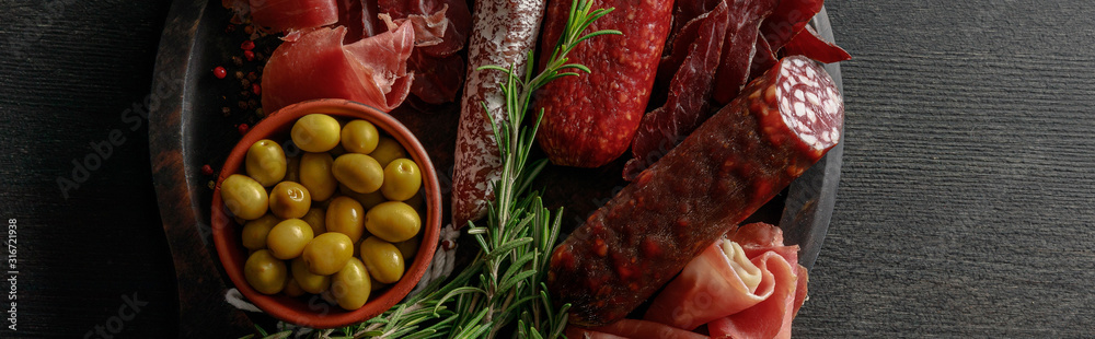 top view of delicious meat platter served with olives and herbs on wooden black table, panoramic shot