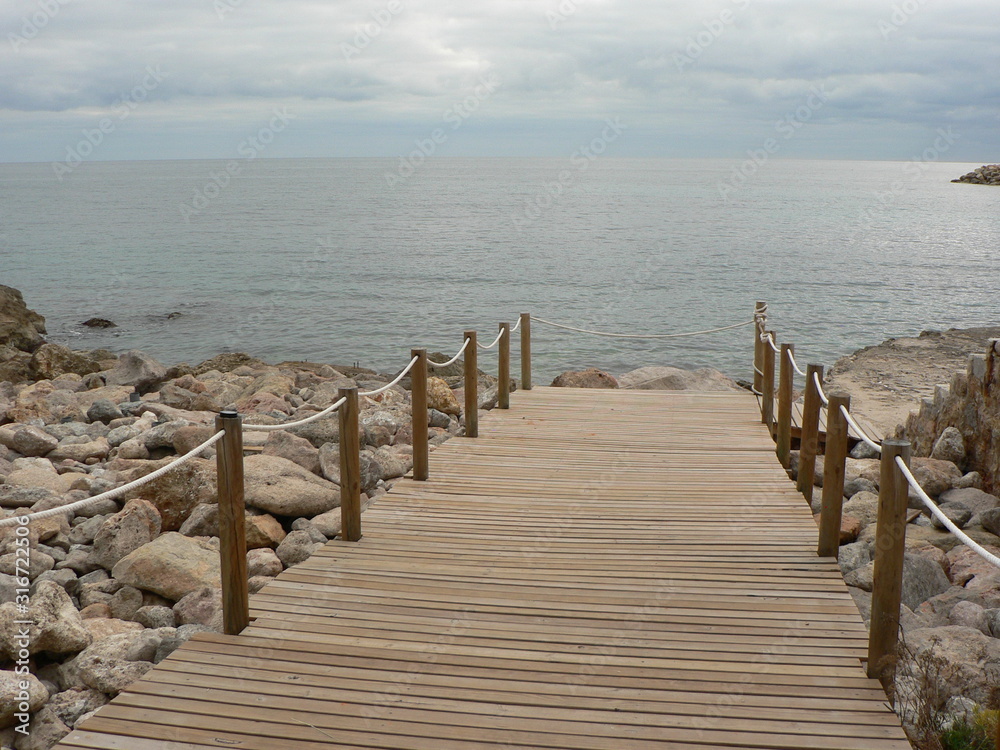 Perspective view of a wooden pier on the coast with sand and blue Mediterranean Sea. Skyline, sea and sky on a cloudy day. Perspective view of a wooden pier on the coast with sand and blue Mediterranean Sea. Skyline, sea and sky on a cloudy day.