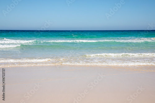 Cloudless sky over a colorful sea with diferent colors aquamarine turquoise dakr blue azure with waves prducing white foam crushing against a yellow beach summer sumertime sunny calm water tranquil