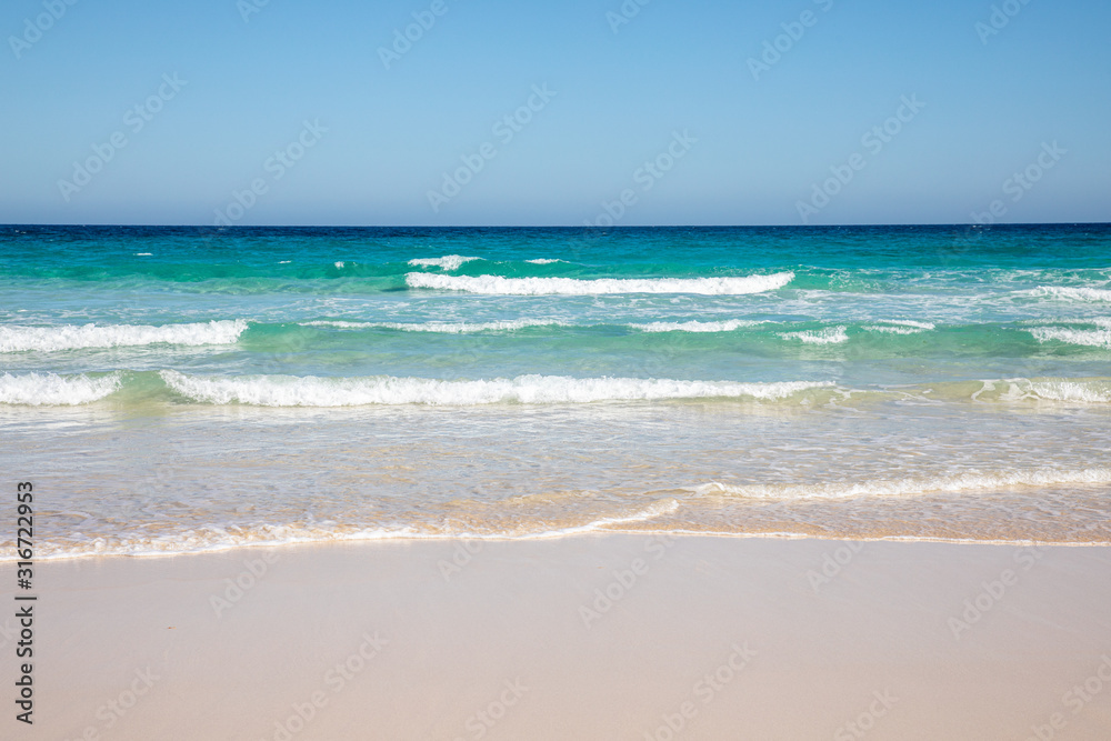 Cloudless sky over a colorful sea with diferent colors aquamarine turquoise dakr blue azure with waves prducing white foam crushing against a yellow beach summer sumertime sunny calm water tranquil
