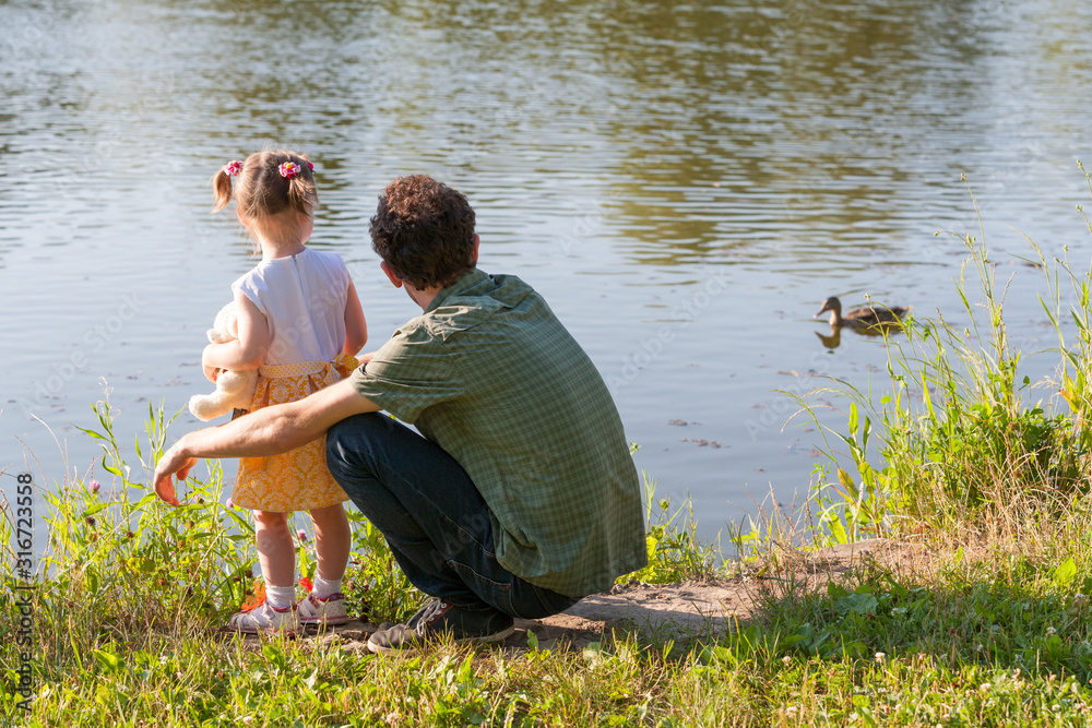Father and daughter looking at lake. Girl is holding a Teddy bear. A wild duck swims through the water. Back view. Father and daughter looking at lake. Girl is holding a Teddy bear. A wild duck swims through the water. Back view.