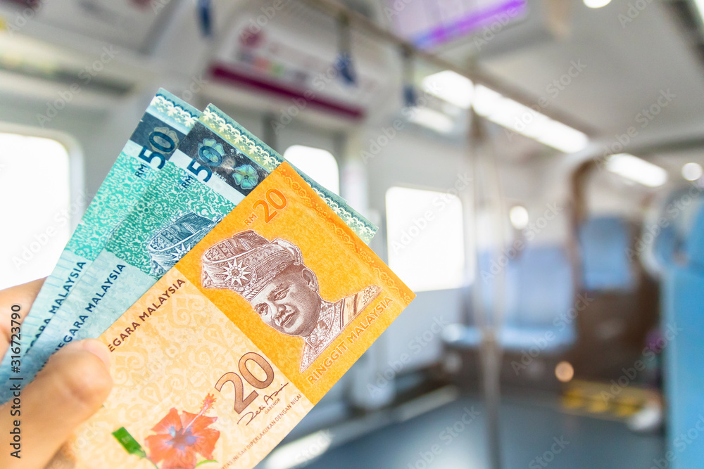 Stack of Ringgit Malaysia bank note on hand and inside KLIA Transit train on background. It is a commuter rail service in Kuala Lumpur, Malaysia between KL Sentral and airport KLIA and KLIA2.