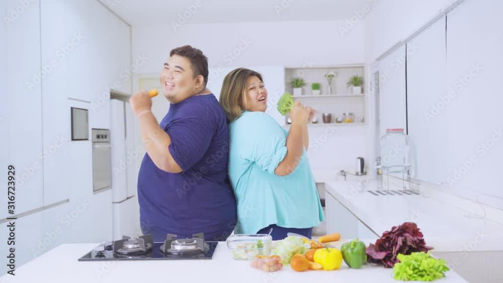 Cheerful overweight couple singing in the kitchen while holding vegetables. Shot in 4k resolution Cheerful overweight couple singing in the kitchen while holding vegetables. Shot in 4k resolution