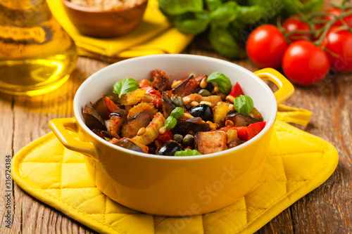 Traditional Italian caponata, served with croutons in a yellow, sunny pot. Decorated with basil and pine nuts.