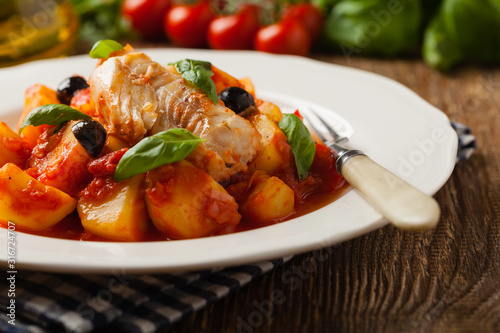 Traditionally prepared cod in Spanish. Served in tomato sauce with boiled potatoes, olivki and basil. Front view. photo