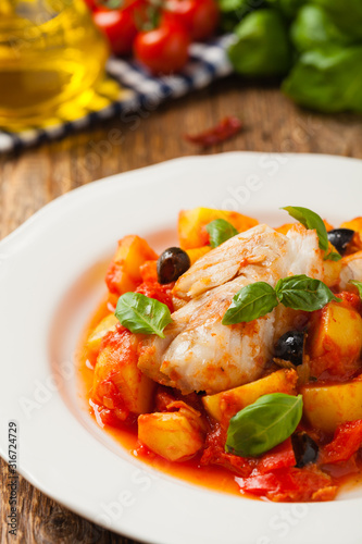 Traditionally prepared cod in Spanish. Served in tomato sauce with boiled potatoes, olivki and basil. Front view.