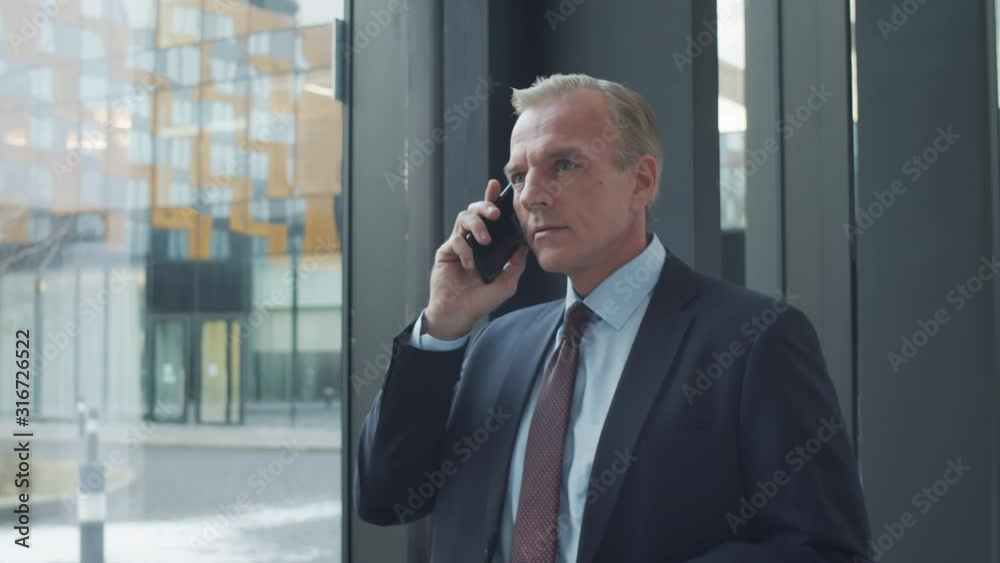 Waist-up arc shot of middle-aged Caucasian businessman in suit and tie standing in hallway of office building next to entrance, looking out into street and talking on mobile phone Waist-up arc shot of middle-aged Caucasian businessman in suit and tie standing in hallway of office building next to entrance, looking out into street and talking on mobile phone