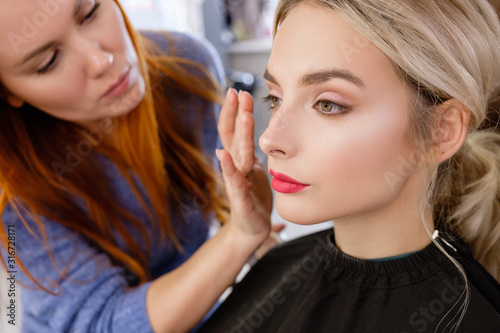 Young red-haired woman make-up artist puts make-up on face of beautiful blonde girl in beauty salon. Concept of personal care and celebration preparation. Daily self-care, beauty treatments in spa