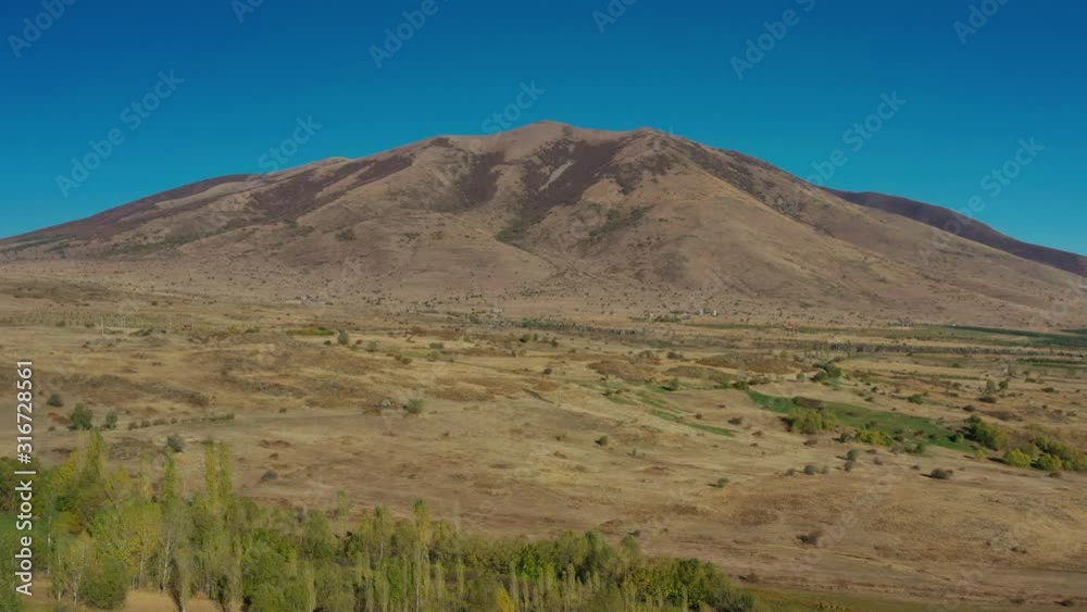 Aerial drone shot fly over zoom out of fields and trees on background of Mount Aragats in summer under birght blue sky. Sandy mountain and green trees in summer time Aerial drone shot fly over zoom out of fields and trees on background of Mount Aragats in summer under birght blue sky. Sandy mountain and green trees in summer time