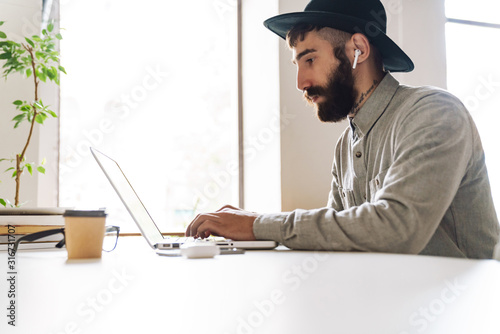 Photo of serious young man using laptop and earpods while sitting