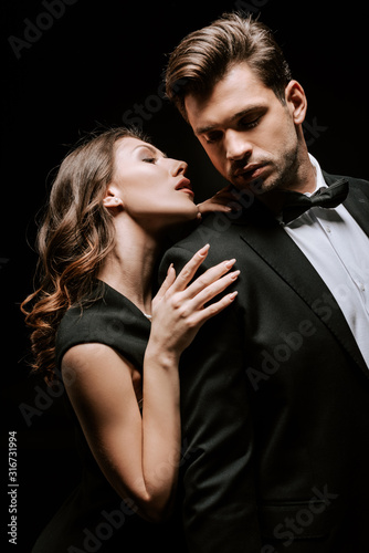 attractive woman touching handsome man in suit isolated on black