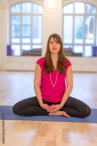 Young woman sitting cross-legged practicing yoga on a mat demonstrating lotus position, asana yoga position