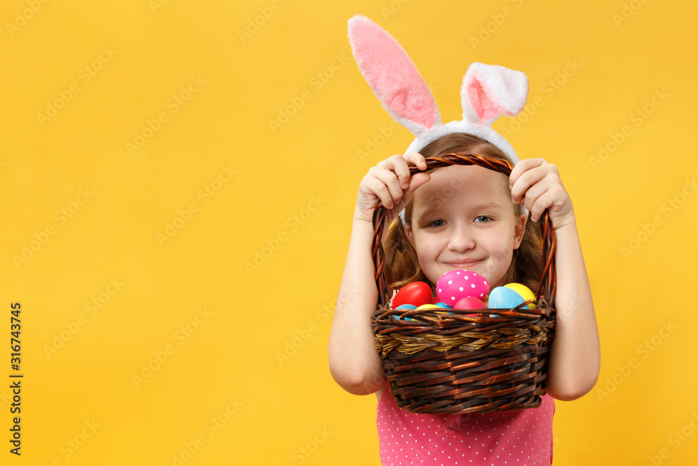 A cheerful child in the Easter ears of a bunny peeks out from under the handle of a basket. Little girls on a yellow background