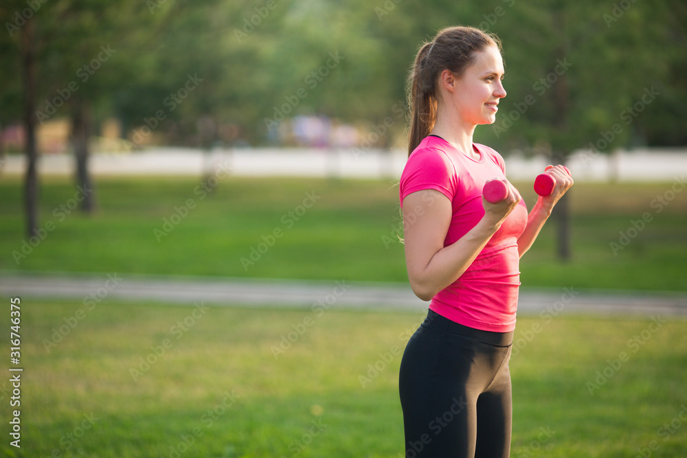 beautiful young woman in sportswear is engaged in training with dumbbells in the summer in the park beautiful young woman in sportswear is engaged in training with dumbbells in the summer in the park