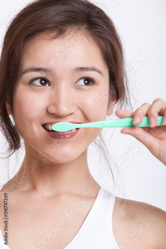 Young Asian woman in white tank top brushing teeth against white background