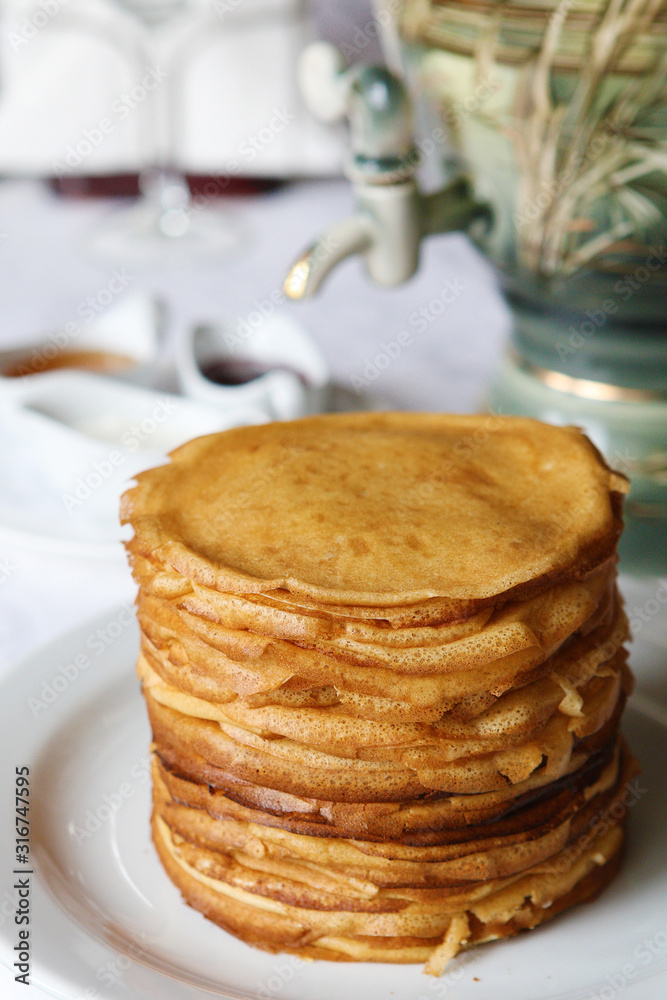 Stack of thin crepes with tea, sour cream, jam and honey. Dish served with a faience samovar on a table covered with a white tablecloth. Stack of thin crepes with tea, sour cream, jam and honey. Dish served with a faience samovar on a table covered with a white tablecloth.