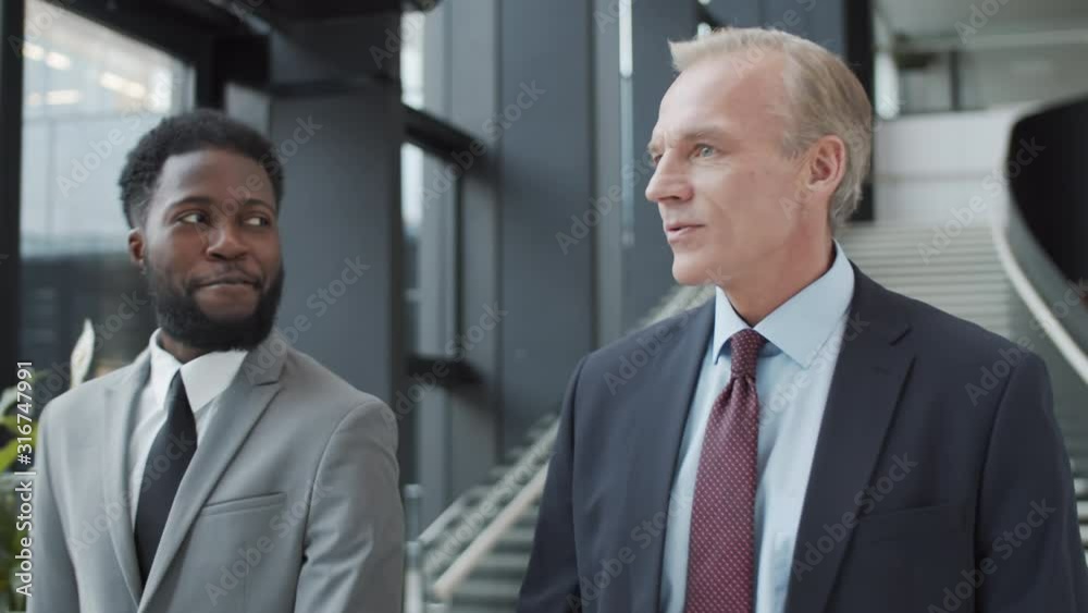 Chest-up tracking shot of two middle-aged Caucasian and young Afro-American businessmen in formal suits walking through hallway of office building, talking politely and smiling Chest-up tracking shot of two middle-aged Caucasian and young Afro-American businessmen in formal suits walking through hallway of office building, talking politely and smiling