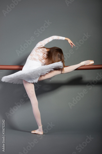 Young female ballet dancer stretching at ballet bar over grey background