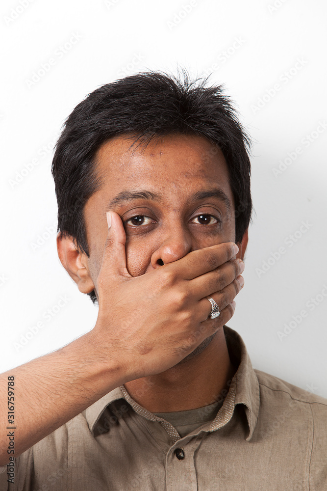 Portrait of young Indian man covering his mouth against white background Portrait of young Indian man covering his mouth against white background