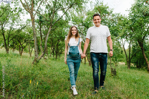 Pregnant girl and her husband are happy to hold hands, goes in the outdoor in the garden background with trees. Close up. full length. looking at camera