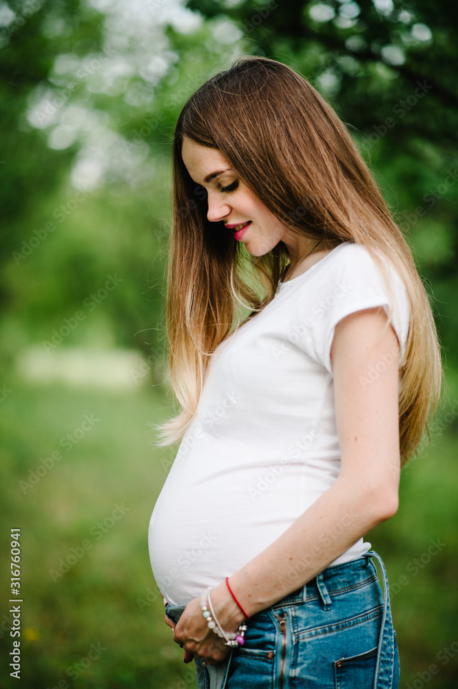 Pregnant happy girl stand and hold hands on stomach, stand in the outdoor in the garden background with green trees. Close up. upper half. Looking down Pregnant happy girl stand and hold hands on stomach, stand in the outdoor in the garden background with green trees. Close up. upper half. Looking down