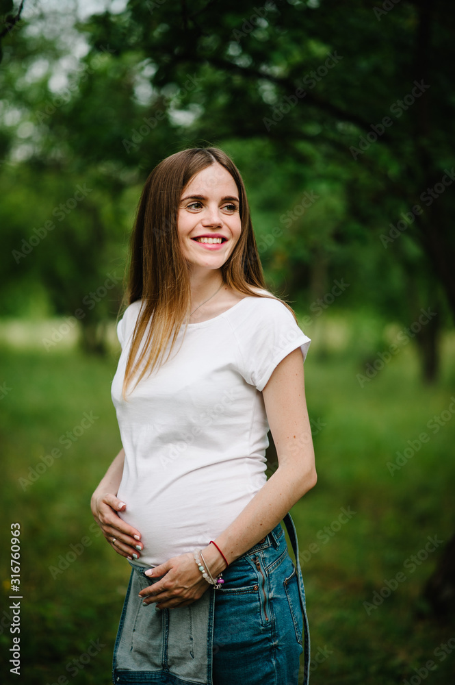 Pregnant happy girl stand and hold hands on stomach, stand in the outdoor in the garden background with green trees. Close up. upper half. half length. Looking sideways. Pregnant happy girl stand and hold hands on stomach, stand in the outdoor in the garden background with green trees. Close up. upper half. half length. Looking sideways.