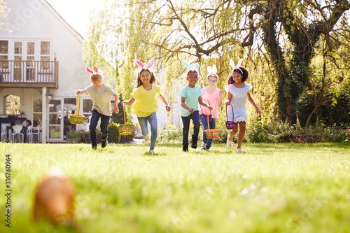 Group Of Children Wearing Bunny Ears Running To Pick Up Chocolate Egg On Easter Egg Hunt In Garden photo