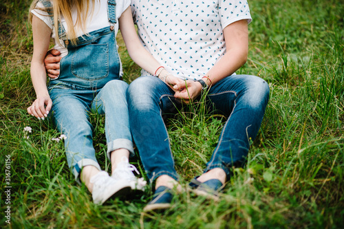 Pregnant girl and her husband to hug, hold hands, on stomach, sitting on the grass in the outdoor in the garden background. Close up. lower half