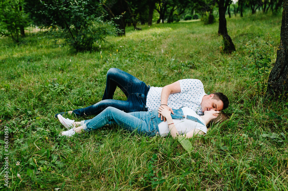 Pregnant girl and her husband are happy to hug, hold hands, on stomach, lie on the grass in the outdoor in the garden background. Close up. full length. Looking at each other and kissing. Side view. Pregnant girl and her husband are happy to hug, hold hands, on stomach, lie on the grass in the outdoor in the garden background. Close up. full length. Looking at each other and kissing. Side view.