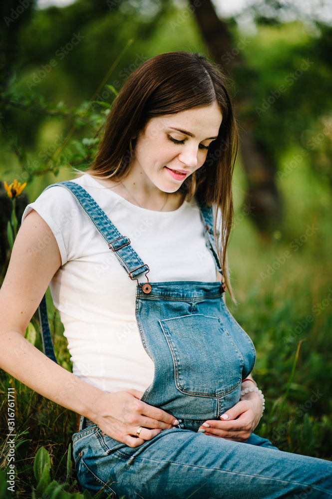 Pregnant beautiful young happy girl, hold hands on stomach, sitting on grass. Close up. upper half. looking down. Pregnant beautiful young happy girl, hold hands on stomach, sitting on grass. Close up. upper half. looking down.