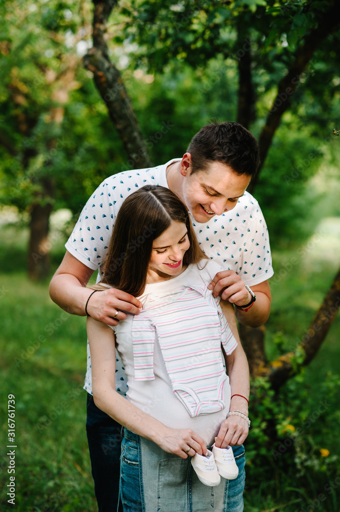 Pregnant girl and husband hold hands to hug, to put baby shoes and clothes on stomach, waiting baby. stand in the outdoor in the garden background. Close up. upper half. looking down Pregnant girl and husband hold hands to hug, to put baby shoes and clothes on stomach, waiting baby. stand in the outdoor in the garden background. Close up. upper half. looking down
