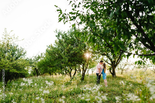 Pregnant girl and her husband to hold hands and kiss, stand in the outdoor in the garden background with trees. full length.