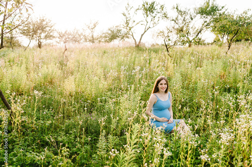 Pregnant happy girl sits and hold hands on stomach,  sitting on grass in field in the outdoor in the garden background with trees. full length. Looking at camera. © Serhii