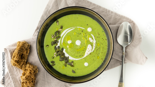 Green broccoli soup with cream, olive oil and pumpkin seeds in a black bowl on a grey napkin. There is some brown bread and spoon near it. White background. 