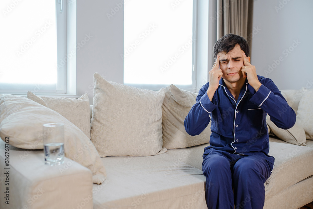 Young caucasian man in bedroom sitting on the sofa and suffer from headache